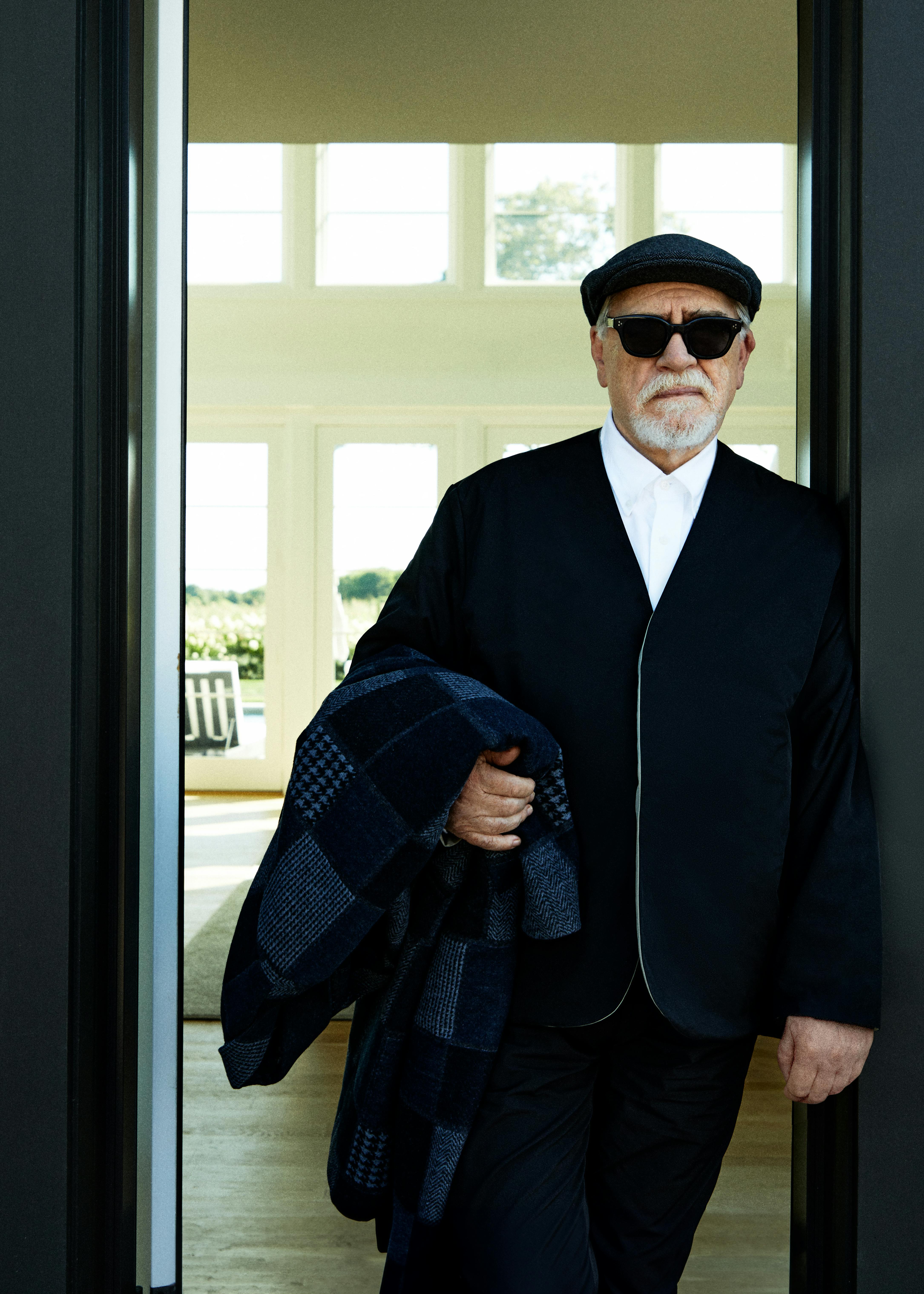 A photograph of Brian Cox, the actor famous for portraying Logan Roy on 'Succession,' wears a black Kith blazer, sunglsases, and white shirt, holding a patchwork coat