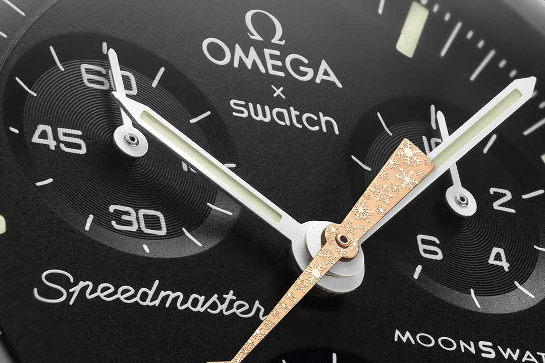 A close-up photo of OMEGA & Swatch's cold moon-inspired Moonswatch watch collab, showing a black watch face and gold hands with a snowflake pattern