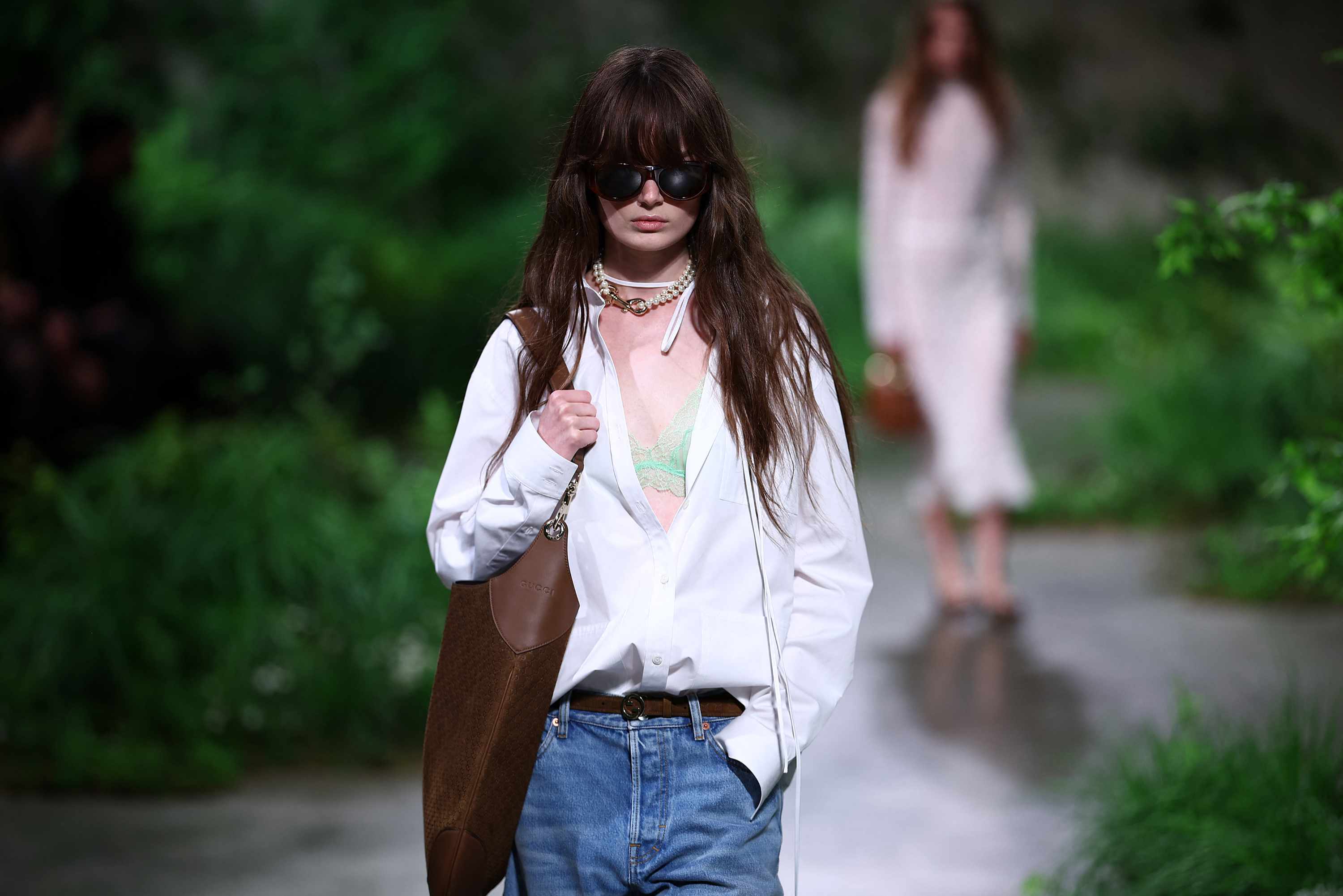 A model wears Gucci Cruise 2025 white shirt bra, jeans, and bag with dark sunglasses