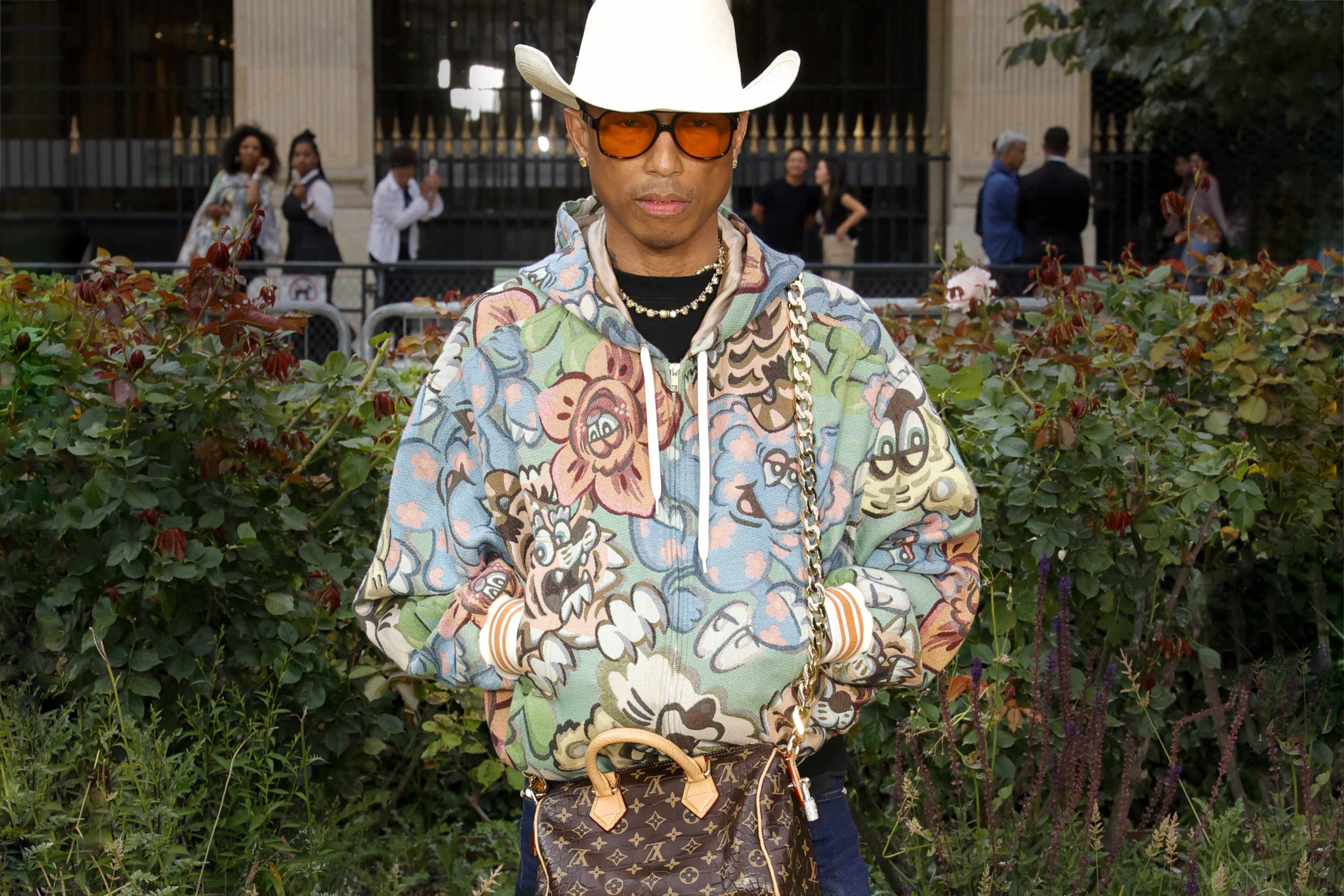 Pharrell wearing a white cowboy hat, patterned jacket, flared blue jeans, adidas sneakers, and $850 louis vuitton millionaires speedy bag