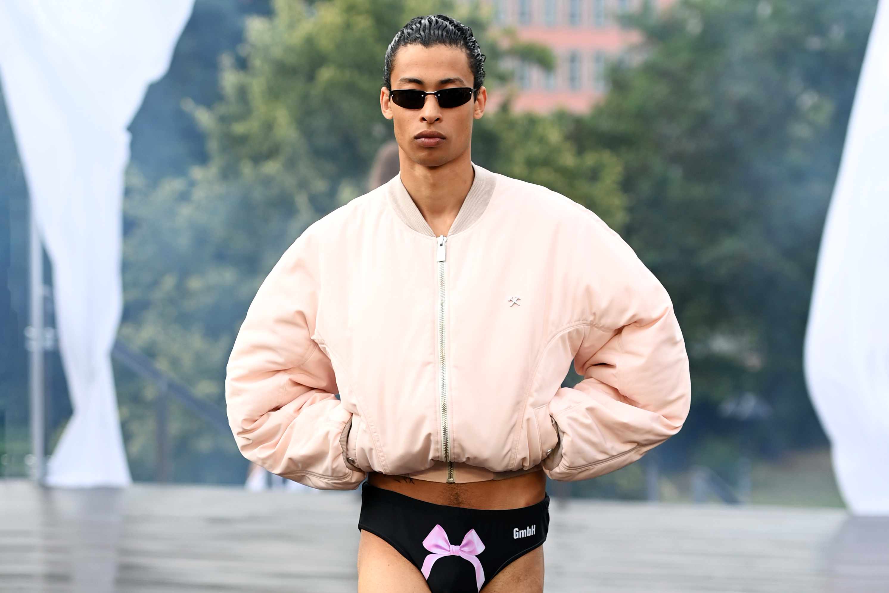 a model wears GmbH's SS25 beige bomber jacket & black underwear with sunglasses and Axel Arigato sneakers