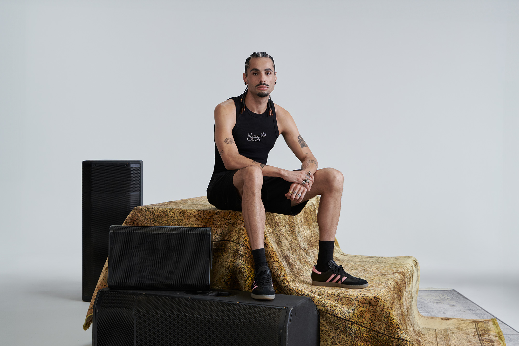 A man wearing a black tank top and shorts sits on a carpet and large speakers