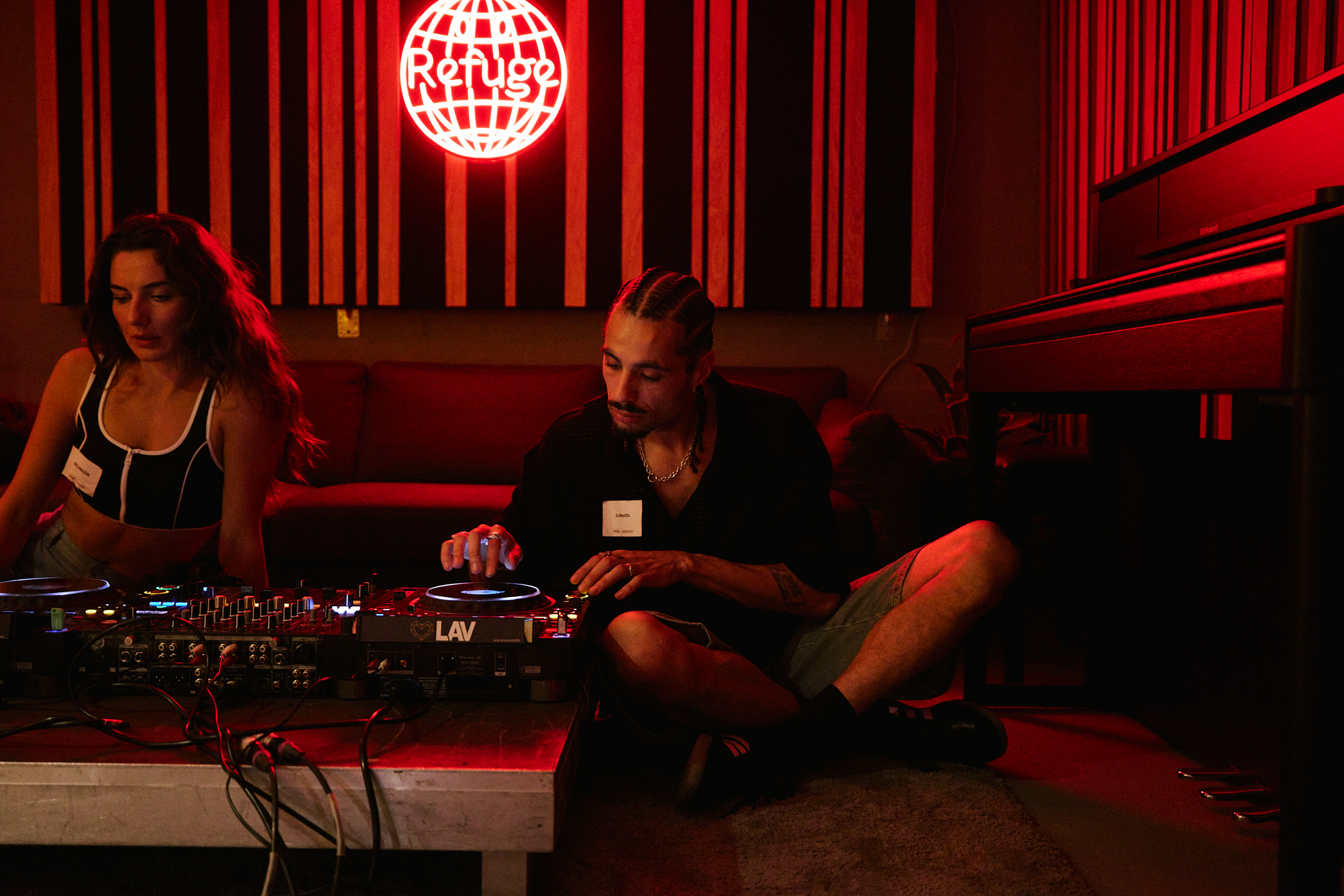 Two DJs sit cross-legged at decks in dim red lighting
