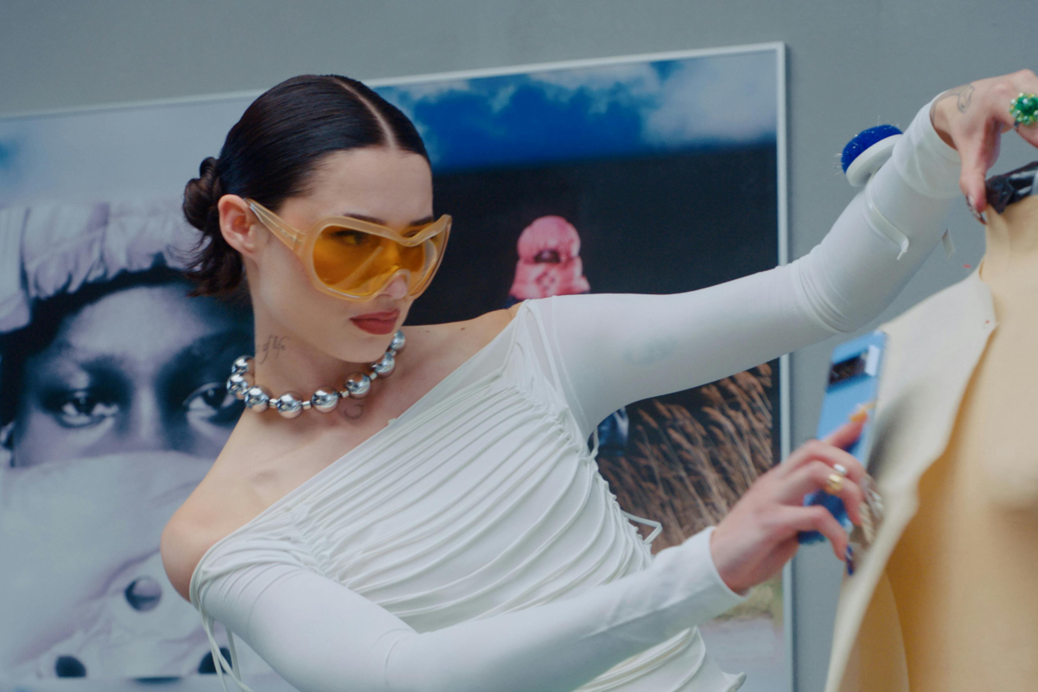 A woman wearing large sunglasses holds a phone against a mannequin