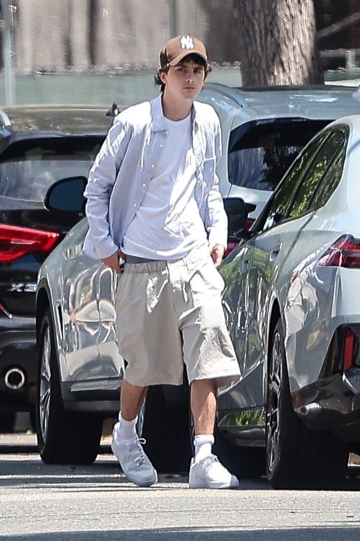 Timothee Chalamet wears a blue shirt, white T-shirt, brown yankees hat, beige shorts and white nike air force 1 sneakers while out in los angeles with his mother